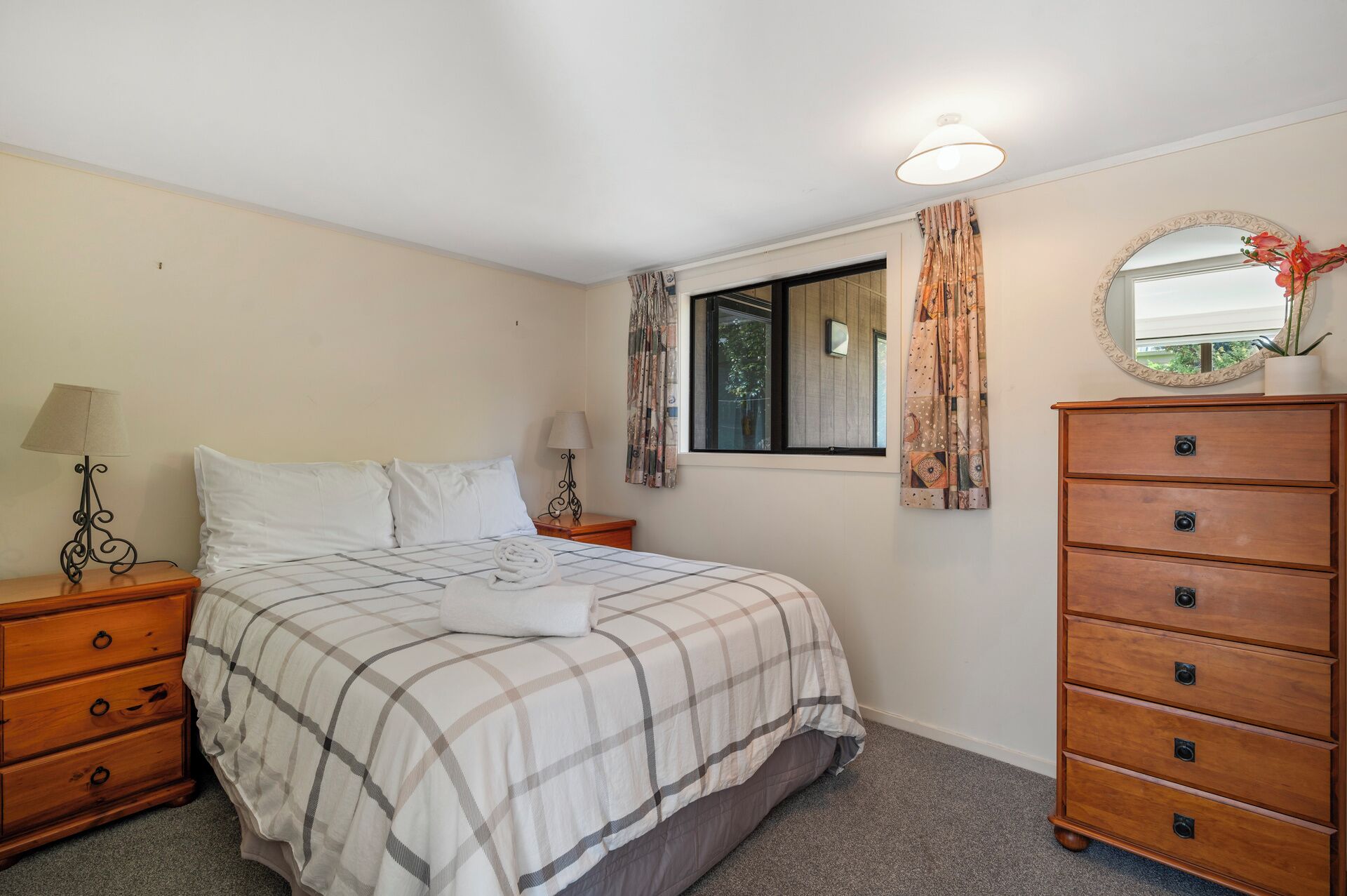 A bedroom with a bed, a dresser, two bedside tables with lamps, a window with curtains, and a mirror on the wall.