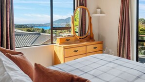 A bedroom with a large bed, a wooden dresser with a mirror, and a view of a cityscape and mountains through the window. - Acacia Views - Acacia Bay Holiday Home (Acacia Bay)