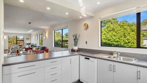 A modern kitchen with white cabinets, a dishwasher, and a large window with a view of trees. - Acacia Views - Acacia Bay Holiday Home (Acacia Bay)