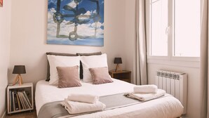 A neatly made bed with white and pink pillows, a bedside table with a lamp, a radiator, and a window with curtains. - Stylish 2 bedroom apart. with breathtaking private Rooftop, garage (Nîmes)