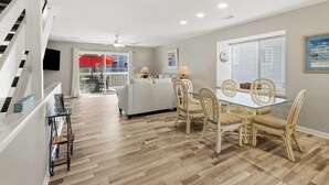 A spacious living area with a glass-top dining table, wicker chairs, and a white sofa. A staircase is visible on the left, and a TV is mounted on the wall. - SEACLUSION OFFERS GULF VIEWS & STEPS TO BEACH! 2 COMMUNITY POOLS! (Port Saint Joe)