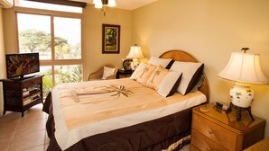 A hotel room with a bed, a ceiling fan, a nightstand with a lamp, a television, and a bookshelf. - Nani Kai Hale #304 on Sugar Beach (Kihei)