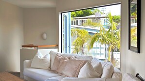 A modern living room with a white sofa, a wooden coffee table, and a large window offering a view of a building and palm trees.