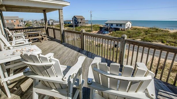 En träveranda med vita Adirondack-stolar, en möblerad sittplats under tak och utsikt över stranden och havet.