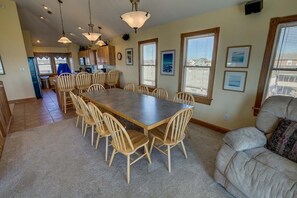 Un grand espace à manger avec une longue table et des chaises, une cuisine en arrière-plan et un coin salon avec un canapé et une fenêtre.