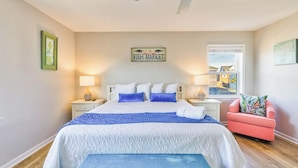 A bedroom with a bed, two bedside tables, a ceiling fan, a window, a pink chair, and a sign on the wall.