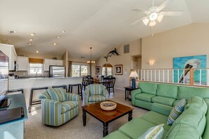 A spacious living room with a green sofa, striped armchairs, and a wooden coffee table. A kitchen is visible in the background with white cabinets and a dining area with chairs.