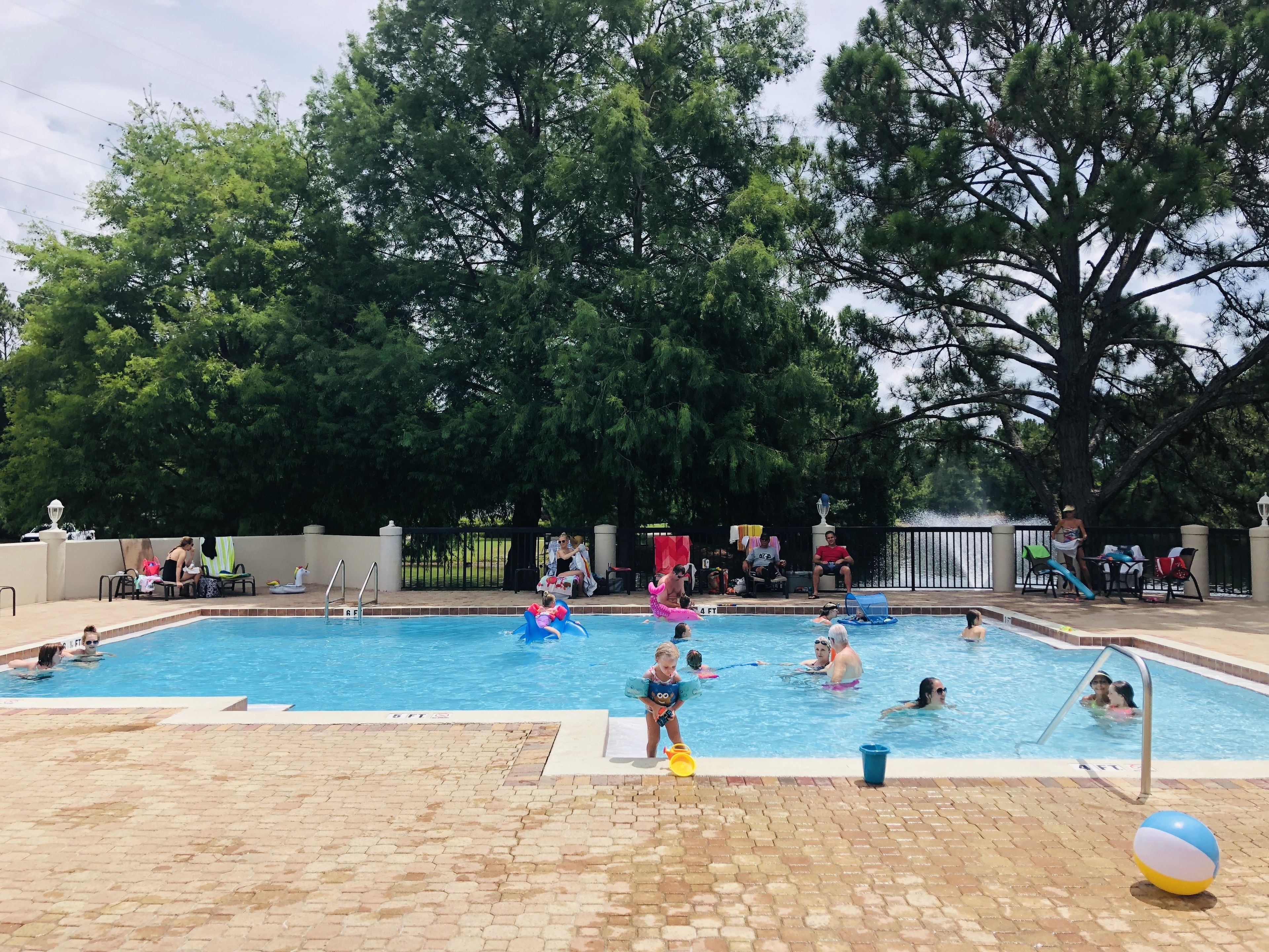Un área de piscina con gente nadando y jugando, rodeada de árboles y una valla.