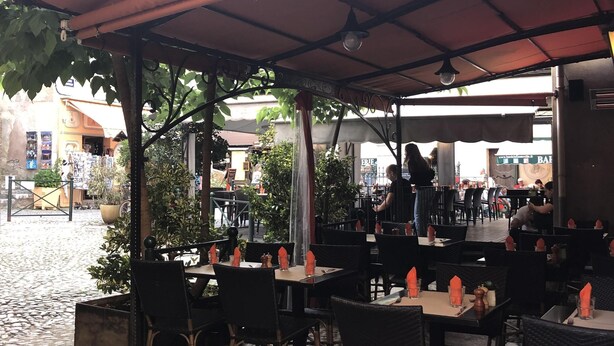 An outdoor cafe with tables and chairs under a covered area, surrounded by greenery and buildings in the background. - Studio in historic district of Corte (Corte)