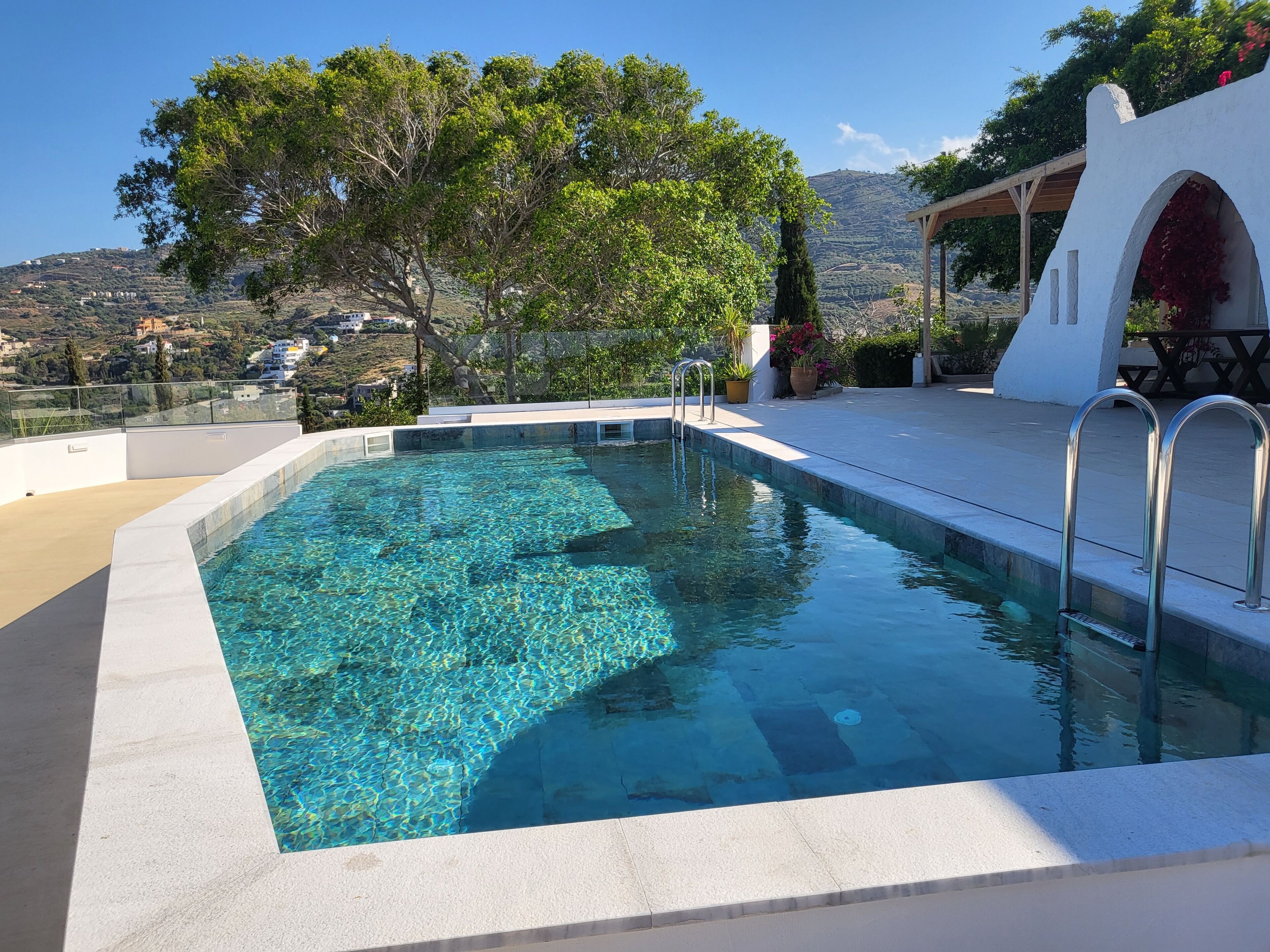 A clear swimming pool with a view of a hillside and a white building with arches.