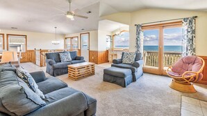 A spacious living room with a large sofa set, a wooden coffee table, and a wicker chair. There is a balcony with a view of the ocean. - Sea Spray | 80 ft from the beach | Dog Friendly, Private Pool, Hot Tub | Kitty Hawk (Kitty Hawk)