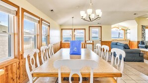 A dining area with a long wooden table and white chairs, a chandelier, and large windows with views of the sea. - Sea Spray | 80 ft from the beach | Dog Friendly, Private Pool, Hot Tub | Kitty Hawk (Kitty Hawk)