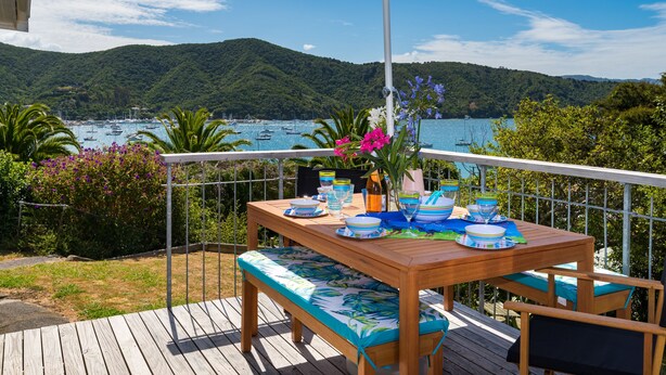 Une terrasse en bois avec une table dressée pour le dîner, offrant une vue sur une baie où se trouvent des bateaux et des collines.