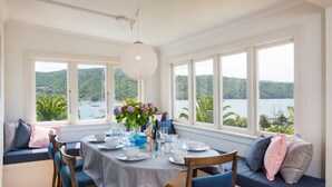 Un coin repas avec une table dressée pour quatre personnes, de grandes fenêtres offrant une vue sur un lac et des collines, et un espace salon confortable avec des coussins.