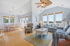 A spacious living room with a high ceiling, large windows, a dining area with white chairs, and a patterned armchair.