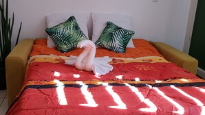 A bed with a red and orange patterned bedspread, two green leaf-patterned pillows, and a white towel. - Studio any comfort. (Bas de la Rivière)