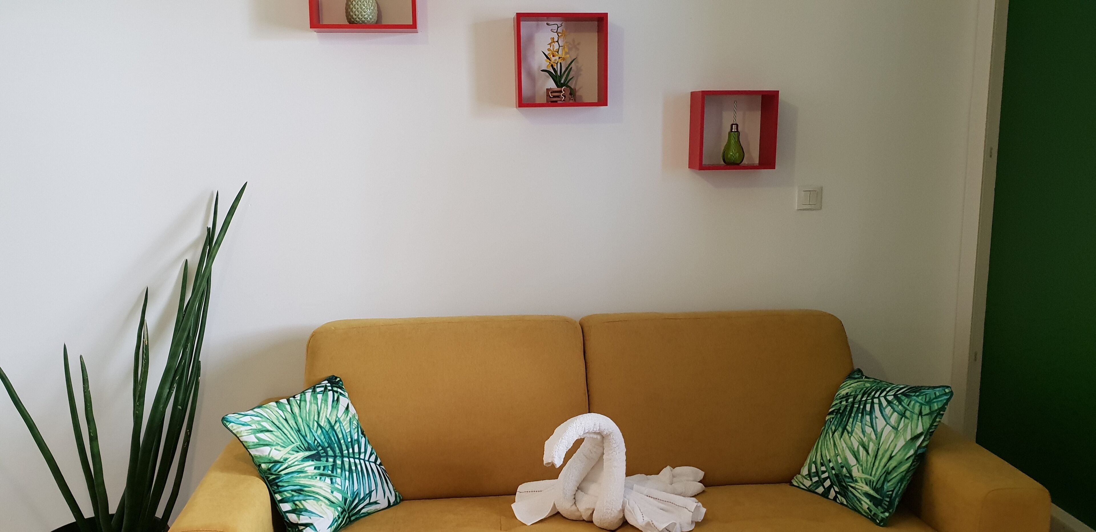 Un canapé jaune agrémenté de deux coussins à motifs de feuilles vertes, d’un coussin blanc en forme de cygne et d’une grande plante verte placée à gauche.