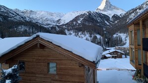 Una cabaña de madera con techo nevado, frente a montañas cubiertas de nieve y un cielo despejado.