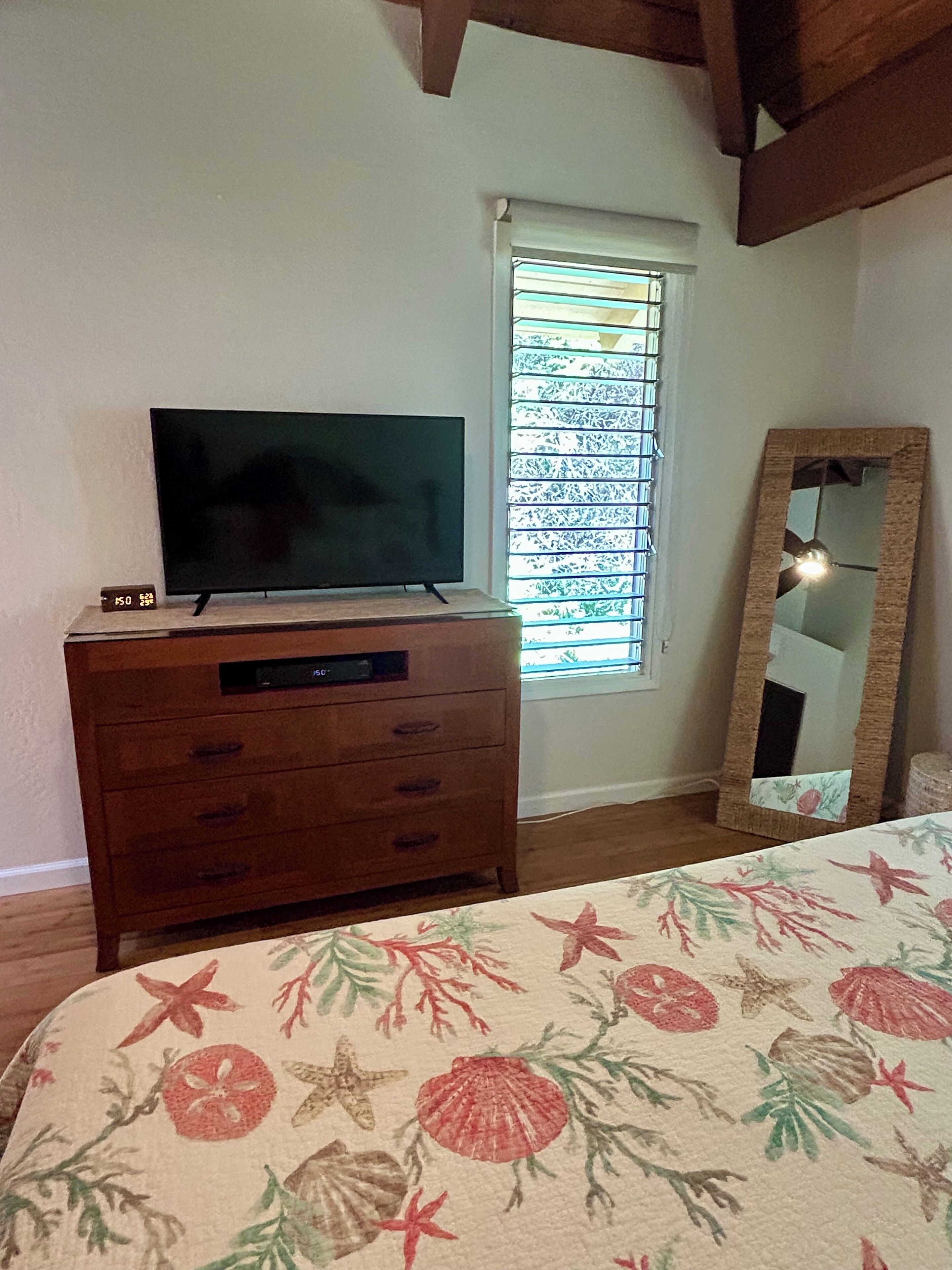 A bedroom with a wooden dresser, a television on top, a window with blinds, a mirror, and a bed with a floral bedspread.