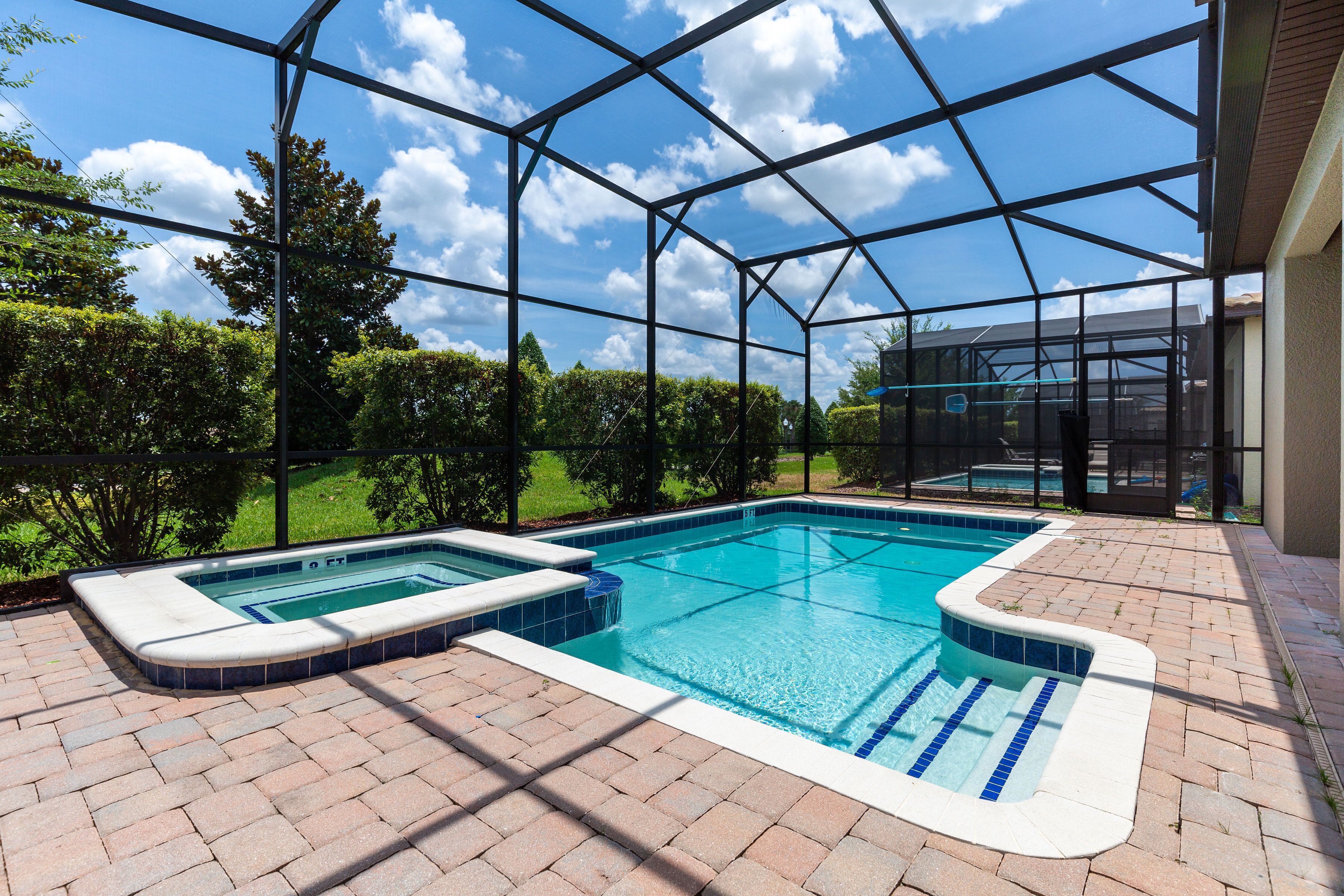 A swimming pool with a glass enclosure, surrounded by a brick patio and a view of a house and greenery.