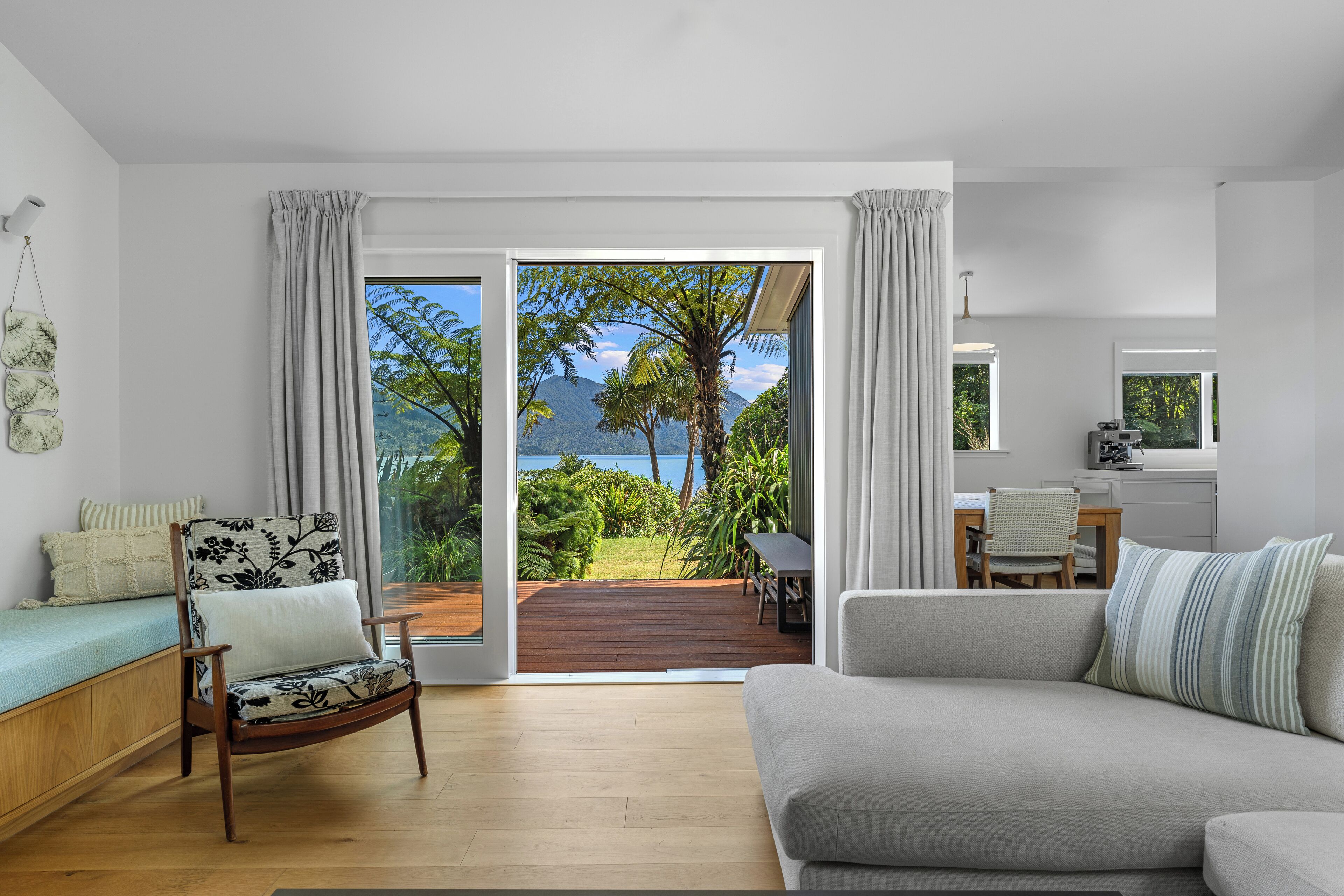 Ett modernt vardagsrum med en soffa, en fåtölj och utsikt över en frodig trädgård och berg genom stora skjutdörrar.