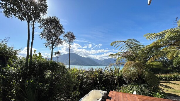 Una terraza de madera con una bañera, rodeada de abundante vegetación y vistas a un lago con montañas al fondo.