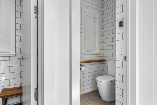A bathroom with white subway tiles, a toilet, a wooden shelf, and a window with blinds.