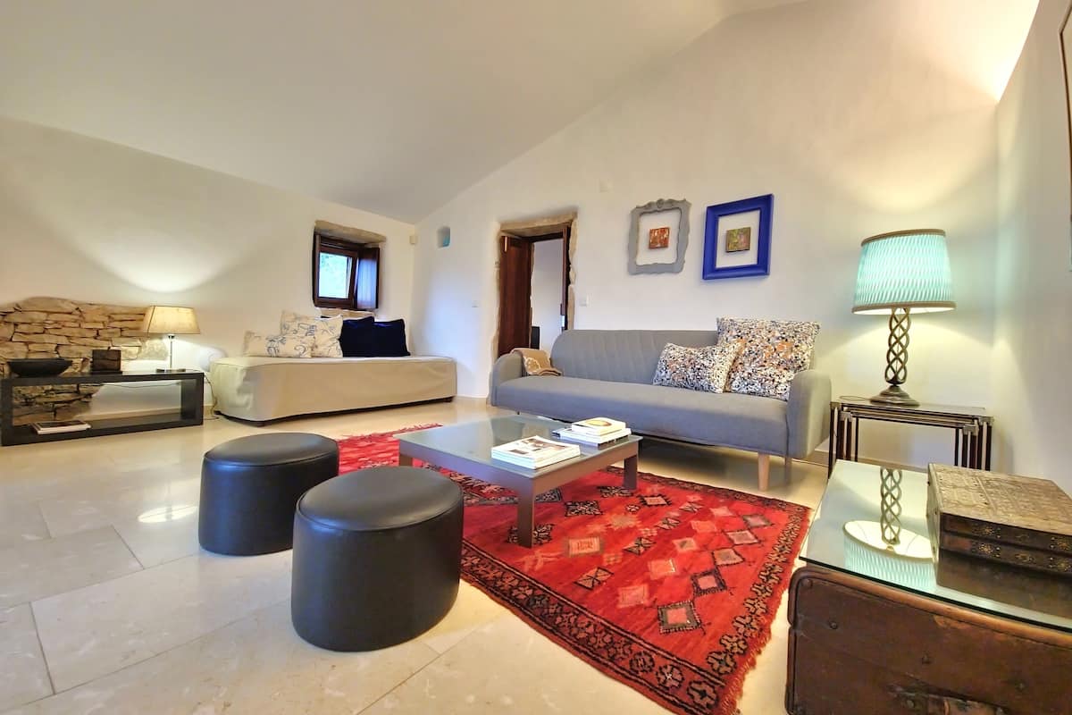 A spacious living room with a stone wall, a grey sofa, a red patterned rug, and a coffee table with books.