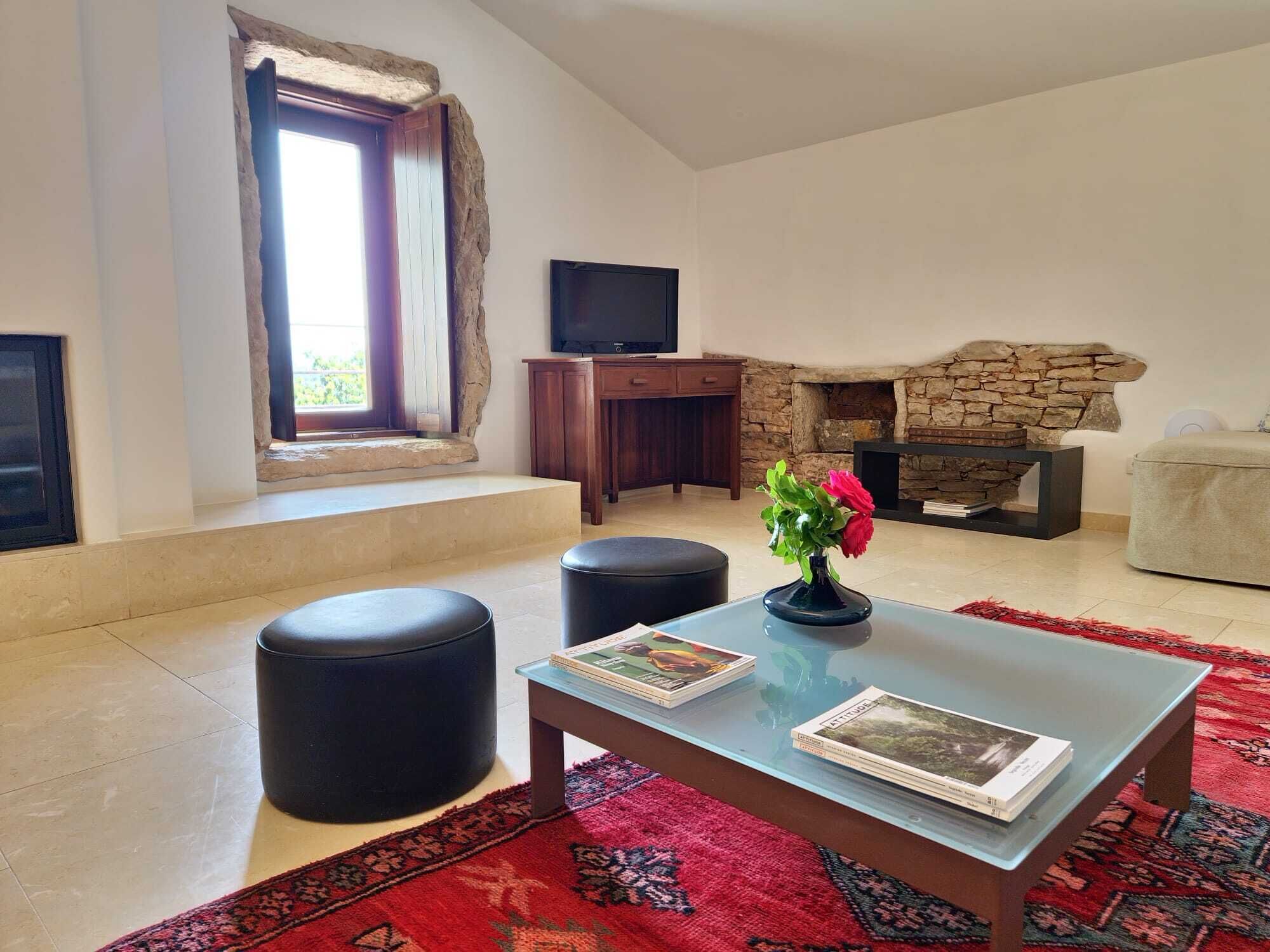 Una sala de estar con chimenea de piedra, un televisor de pantalla plana, una mesa de centro con revistas y un tapete rojo estampado.
