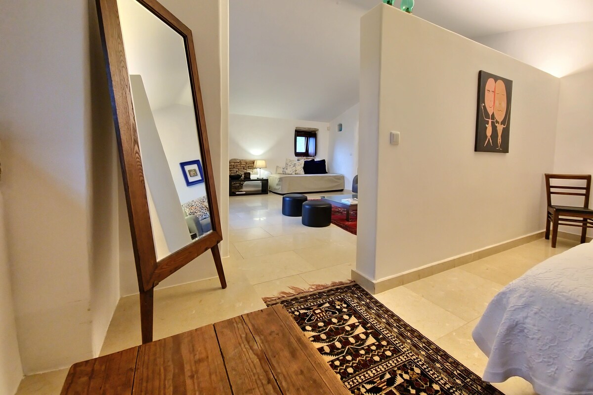 A bedroom with a bed, a wooden table, a mirror, a rug, a chair, and a painting on the wall.