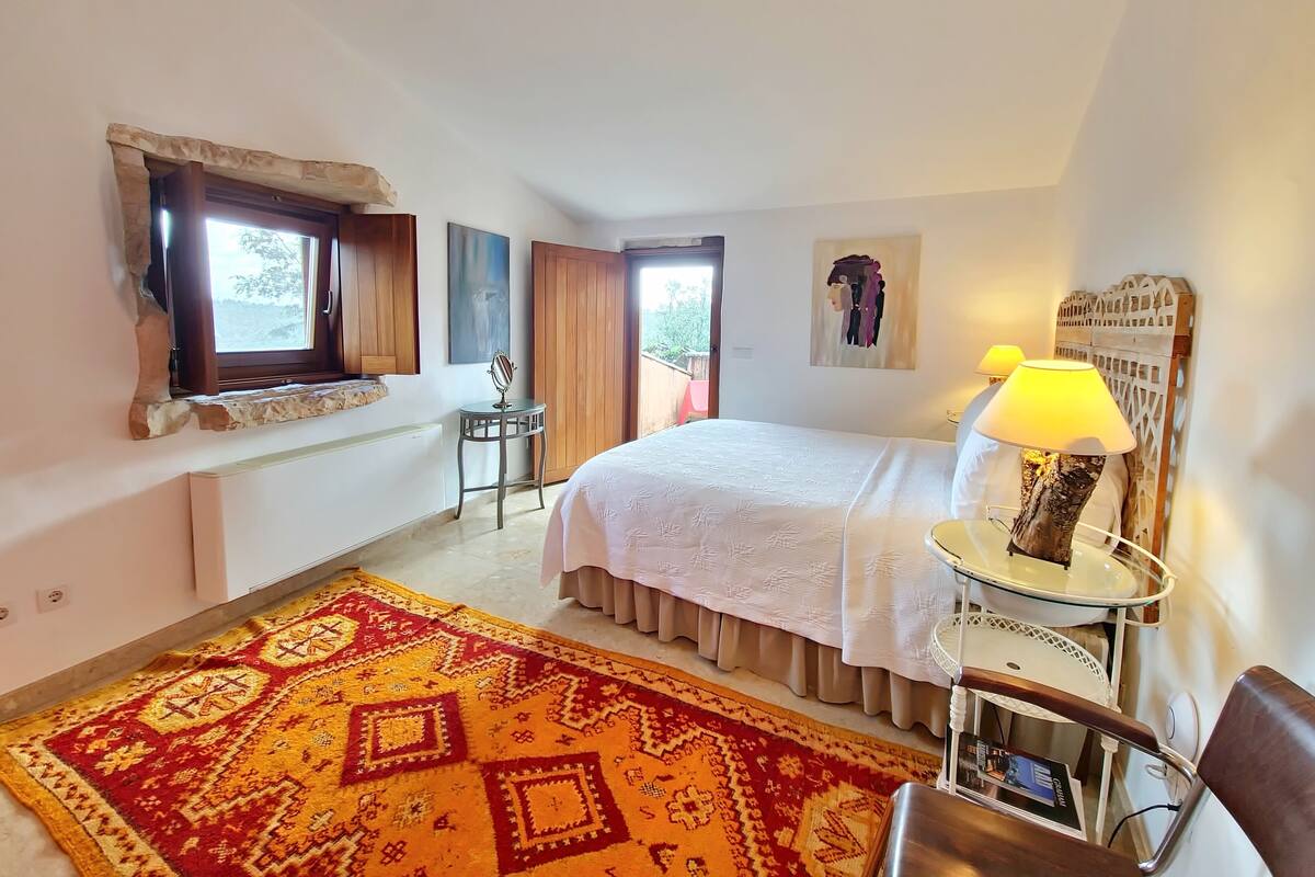 A bedroom with a bed, a bedside table with a lamp, a chair, a window, and a patterned rug.