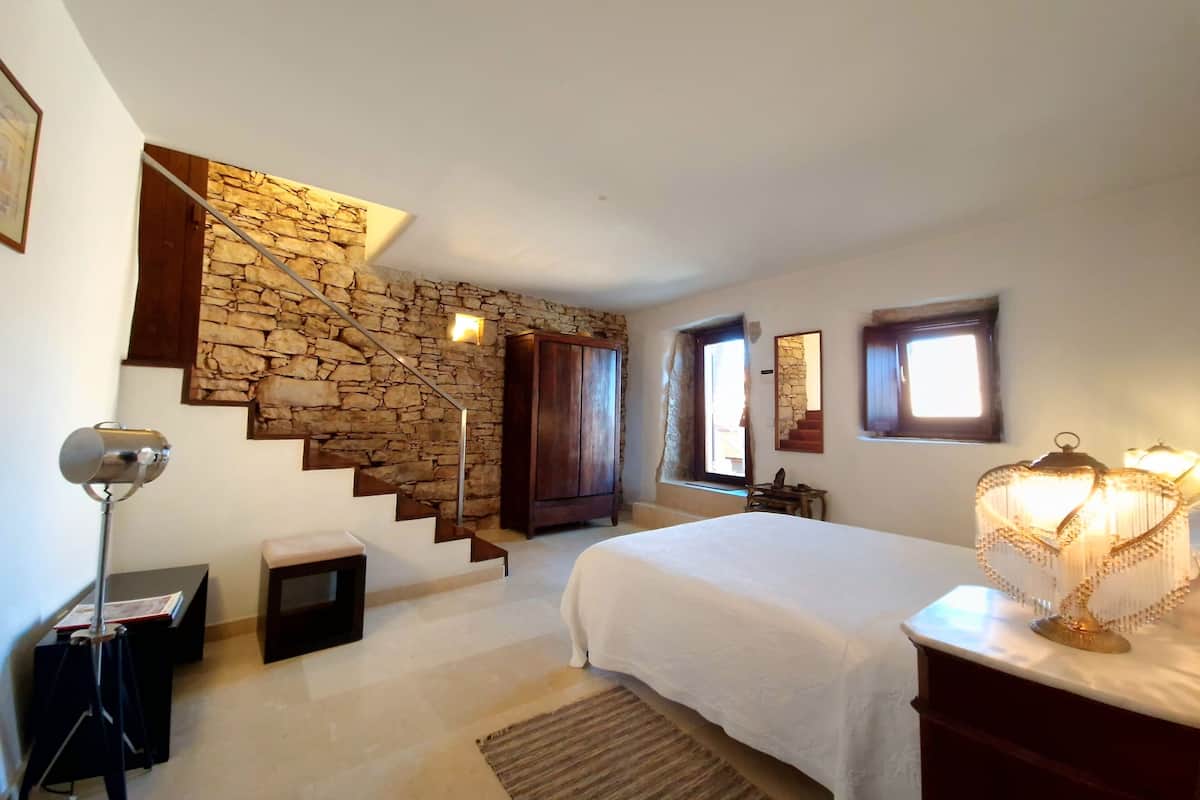 A bedroom with a stone wall, wooden staircase, a bed with a white bedspread, a desk with a lamp, and a window with a view.