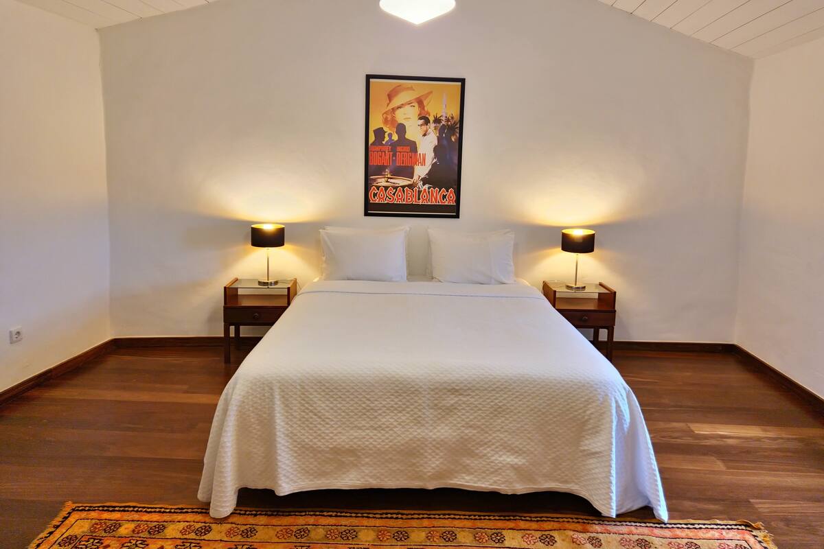 A bedroom with a large bed, two bedside tables with lamps, a poster on the wall, and a patterned rug.