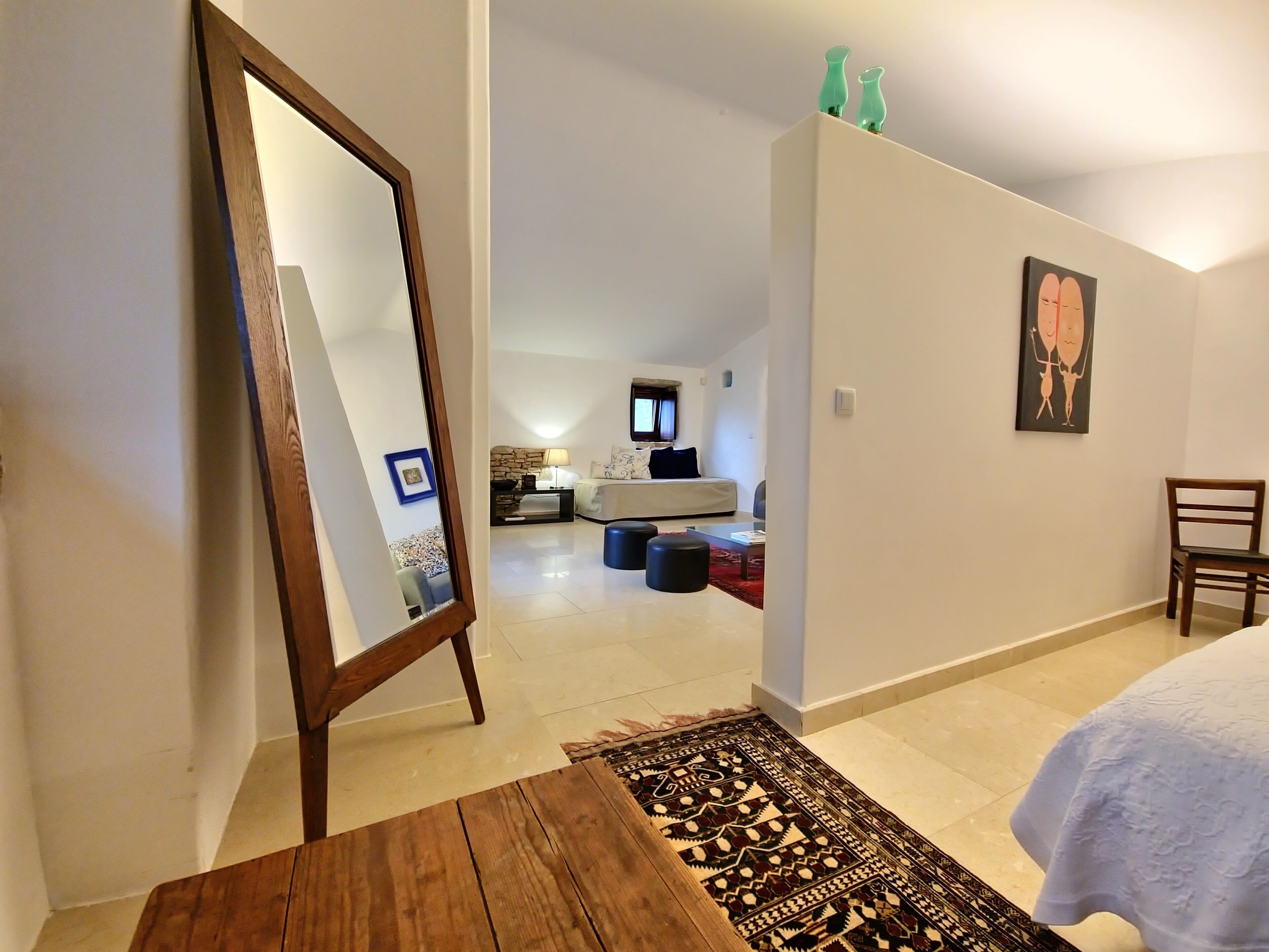 A bedroom with a bed, a wooden table, a mirror, a rug, a chair, and a painting on the wall.