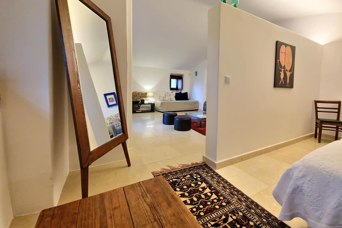 A bedroom with a bed, a wooden table, a mirror, a rug, a chair, and a painting on the wall.