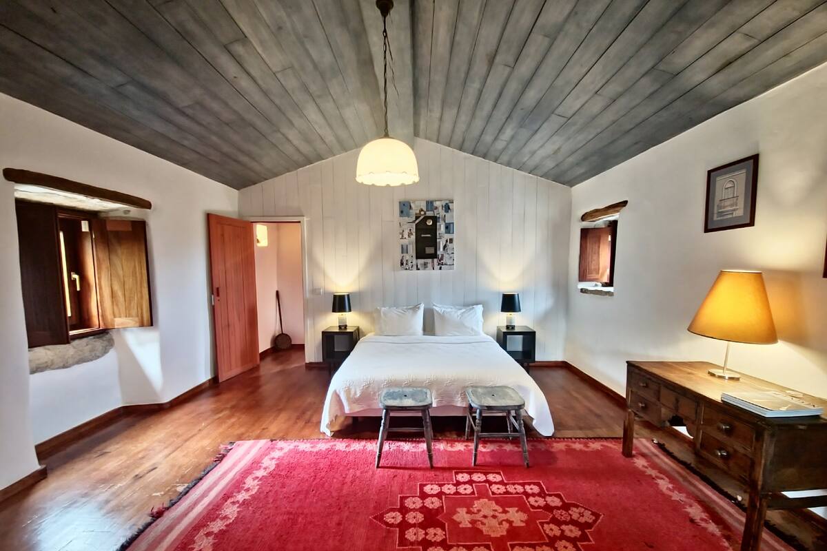 A bedroom with a wooden ceiling, a bed with white linens, two bedside tables with lamps, a red patterned rug, and a wooden dresser.