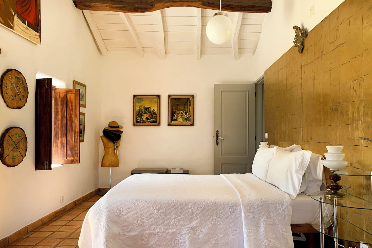 A bedroom with a bed, wooden ceiling beams, a door, and two framed paintings on the wall.