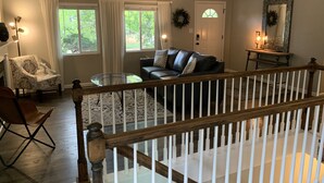 A spacious living room with a leather sofa, a patterned armchair, and a glass coffee table. A wooden railing separates the living area from a staircase.