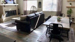 A living room with a fireplace, a black leather sofa, a dining table with chairs, and large windows with curtains.