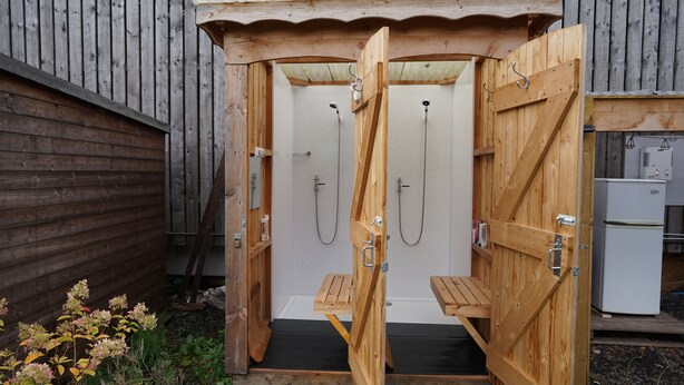 Une cabine de douche extérieure en bois, dotée de deux pommes de douche, d’un banc et d’un petit réfrigérateur.