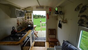 Une cuisine compacte équipée d’une cuisinière à gaz, d’une bouilloire et de divers ustensiles. Une petite table et une chaise en bois se trouvent près de l’entrée, donnant accès à une terrasse.
