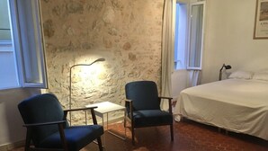 Ein Zimmer mit Steingemäuer, zwei Stühlen, einem kleinen Tisch, einem Bett und einer Lampe.