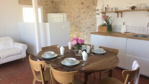 Ein Essbereich mit einem runden Holztisch, gedeckt für vier Personen, umgeben von Holzstühlen. Im Hintergrund ist eine weiße Couch zu sehen, und zur Rechten befindet sich eine Küchenzeile mit einer Holz Arbeitsplatte und verschiedenen Gegenständen.