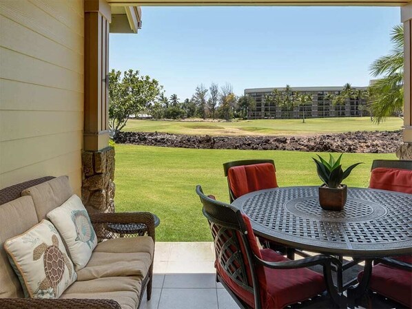 Un patio cubierto con una mesa redonda y sillas, un sofá con cojines, y una vista a un campo de golf y un edificio de hotel al fondo.