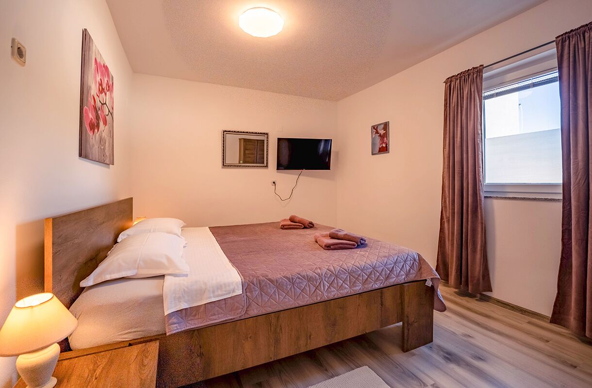 A bedroom with a wooden bed, a flat-screen TV, a window with curtains, and a painting on the wall.