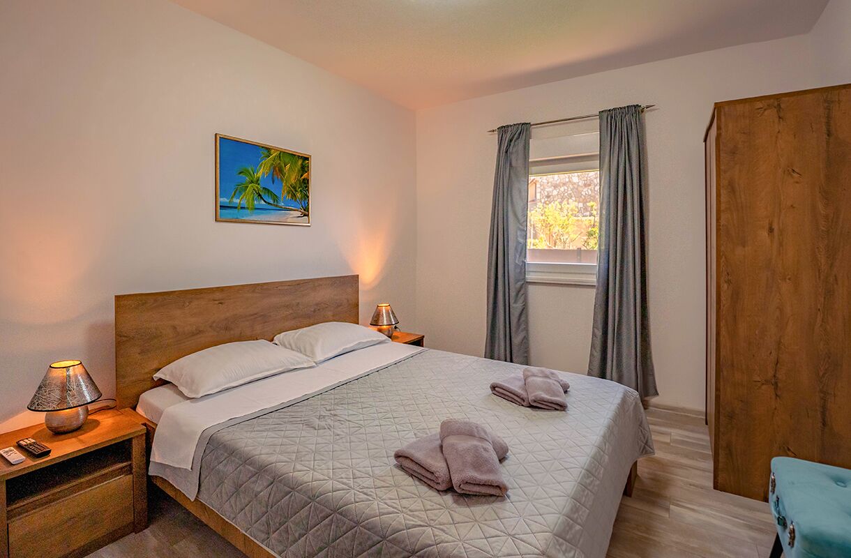 A bedroom with a bed, two bedside tables, a nightstand, a window with curtains, a wooden wardrobe, and a painting of a tropical scene.