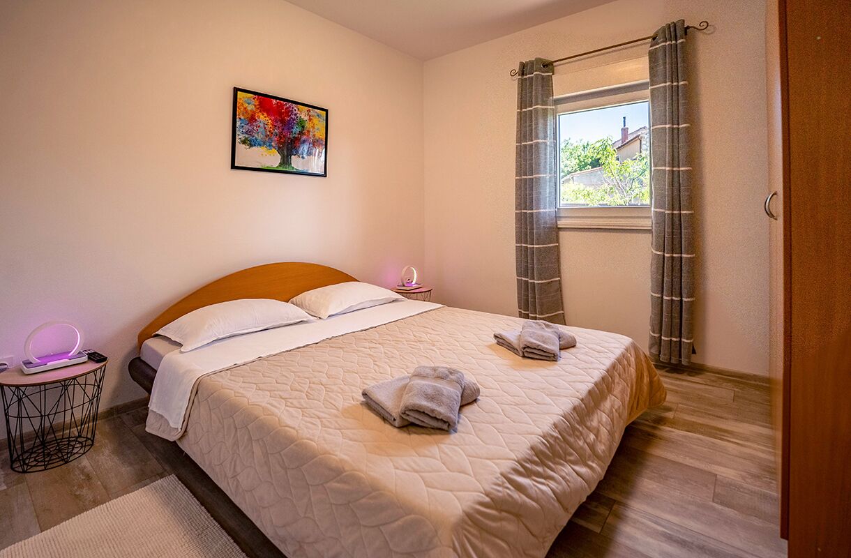 Una cama bien hecha con dos almohadas y una colcha, una mesita de noche con lámpara, una ventana con cortinas y un cuadro colorido en la pared.