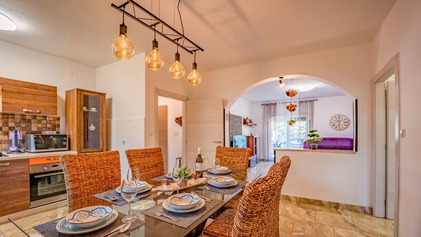 Un coin repas avec une longue table dressée pour quatre personnes, entourée de chaises en osier. Des luminaires sont suspendus au-dessus de la table et une horloge est accrochée au mur.