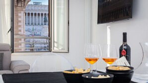 A dining area with a table set for two, two glasses of wine, a bottle of wine, and bowls of snacks. A window reveals a view of a historic building with columns and statues. - Rental In Rome Piazza Venezia View Luxury Apartments (Rome)
