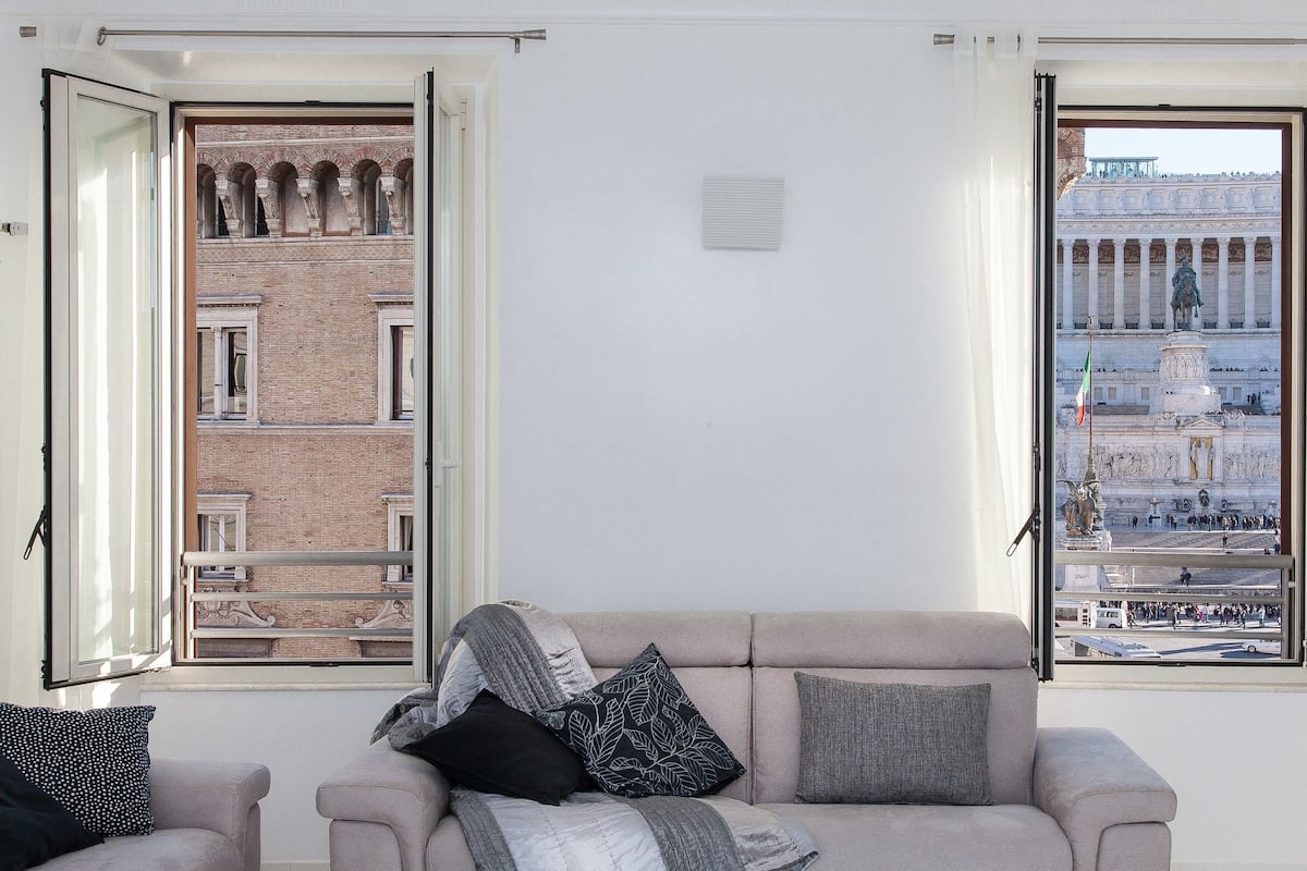 A room with a sofa and two open windows. One window shows a building with arches, and the other shows a large, classical building with columns.